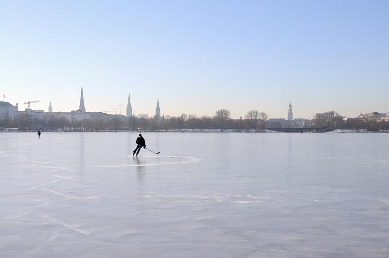 Hamburger Winter - frostiges Winterwetter, Sonnenschein. Hamburger Fotografie.  242_5558 Ein Eishockeyspieler dreht einsam seine Kurven auf dem Alstereis. Im Hintergrund das Panorama von Hamburg mit den Trmen der Hansestadt.